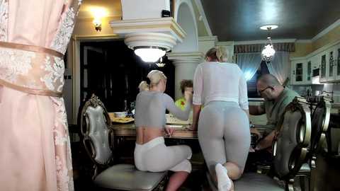 Media: Video: A blonde woman in white pajamas leans over a dining table, while two men in casual attire observe her.