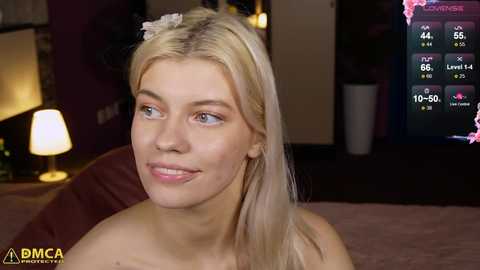 Media: Video of a smiling, fair-skinned blonde woman with straight hair, wearing a pink flower headband, sitting on a bed with a maroon pillow, in a dimly-lit room with a TV showing a game score.