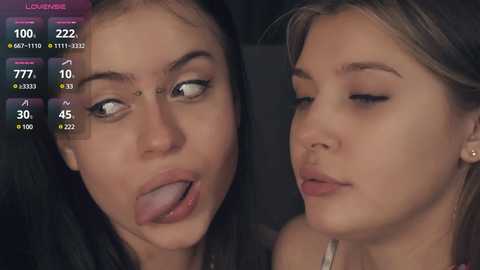 Media: Video of two young women with fair skin, one with dark hair, tongue out, and the other with blonde hair, both with neutral expressions, set against a dark background.