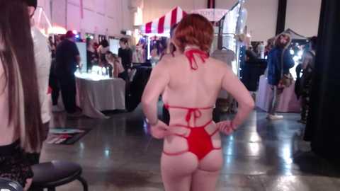 Media: Video of a fair with a woman in a red, strappy bikini, red headband, and hands on hips, walking away. Background includes people, tables, and vendors.