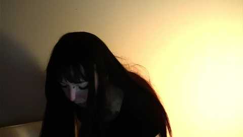 Media: Video of a person with long, dark hair, wearing a white face mask, standing in a dimly lit room with a warm, yellow light source on the right. The person's face is not visible, emphasizing the mask.