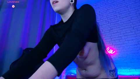 Media: A video shows a light-skinned person with a black long-sleeve shirt, cropped at the waist, and a tattoo on their side, standing against a brick wall with blue and purple lighting.