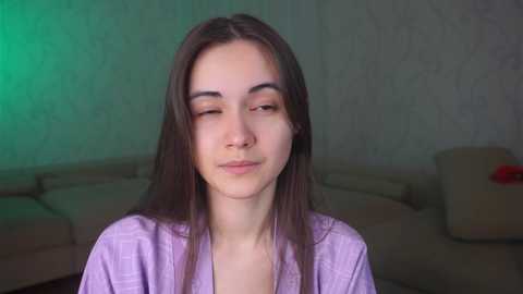 Media: A video of a young woman with long brown hair and fair skin, wearing a light purple robe, sitting indoors with a greenish glow in the background.