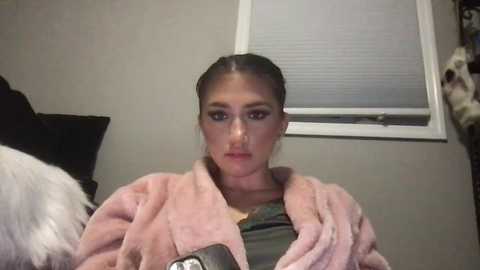 Media: Video of a young woman with fair skin and dark hair tied back, wearing a pink robe over a green shirt, indoors with a window and bed visible.