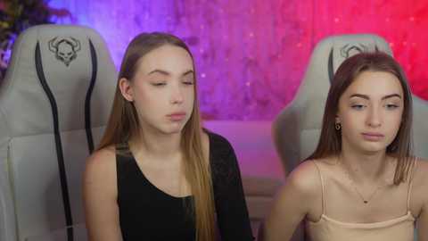 Media: Video of two young women with long hair, one in black, one in beige, seated in gaming chairs, with purple and red lights in the background.