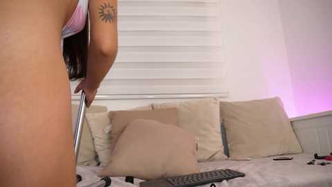 Media: Video of a nude woman with tan skin and long dark hair, holding a pole, standing in a bedroom with beige pillows, a TV remote, and white blinds.