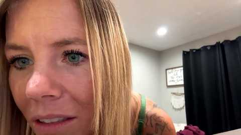 Media: Video of a close-up of a blonde woman with blue eyes and a tattooed arm, wearing a green tank top, in a modern, minimally decorated bedroom with gray walls, a black curtain, and a framed motivational quote.