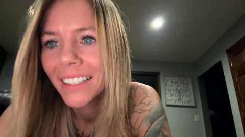 Media: A video of a smiling, light-skinned woman with long blonde hair and blue eyes. She has a tattoo on her left shoulder and is wearing a sleeveless top. The background shows a dimly lit room with gray walls and a wooden door.