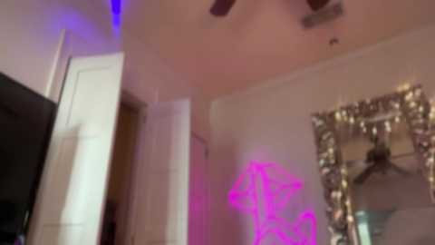 Media: A video of a dimly lit, cozy bedroom with a white wall, a large, ornate mirror, and a purple neon sign emitting a soft glow. A ceiling fan is visible, adding to the room's ambiance.