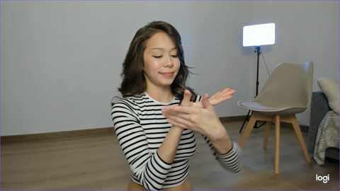 Media: Video of a young Asian woman with wavy black hair, wearing a black-and-white striped long-sleeve top, clapping hands joyfully in a modern, minimalist room with a white wall, wooden floor, and a white chair with a light bulb lamp.