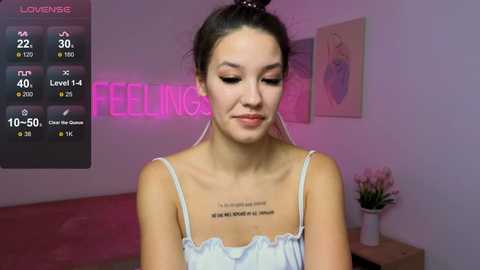 Media: Video of a young woman with dark hair, wearing a light blue camisole, standing indoors with a neon \"Fellings\" sign and flowers in the background.