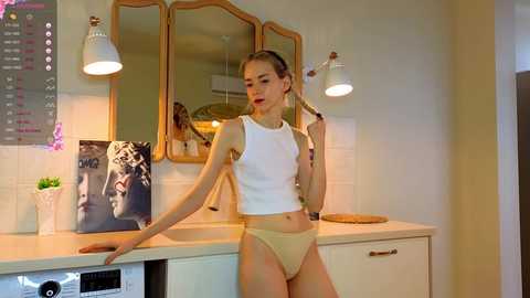 Media: A video of a slender, Caucasian woman with blonde hair in a ponytail, wearing a white crop top and beige panties, leaning against a kitchen counter with white cabinets and a tiled backsplash.