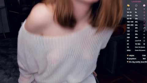 Media: Video of a young, fair-skinned woman with shoulder-length, straight brown hair, wearing a white off-shoulder knit sweater, leaning forward in a dimly lit room.