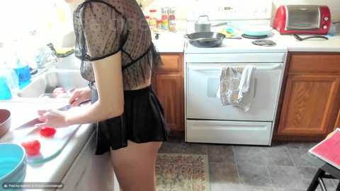 Media: Video of a slender woman with fair skin, wearing a sheer black top and black skirt, cooking in a small, cluttered kitchen with wooden cabinets, a stove, and a sink.