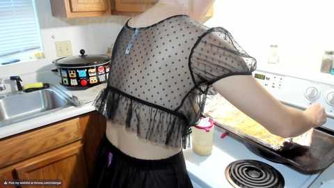 Media: Video of a light-skinned woman with a slender build, wearing a sheer black polka-dotted blouse and black skirt, baking in a kitchen with wooden cabinets and a stainless steel sink.