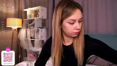 Media: Video of a young woman with long blonde hair, wearing a black top, sitting on a bed. The background features a white shelving unit with clothes, a lamp, and a soft light.