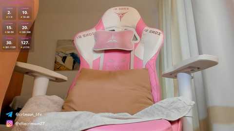 Media: A video of a pink gaming chair with a brown cushion, white armrests, and a pink headset, set in a room with white curtains and a wooden shelf.