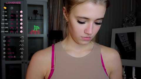 Media: Video of a young Caucasian woman with fair skin, light brown hair, wearing a pink and beige tank top, standing indoors with dim lighting. Background includes a bookshelf, a television, and a pink neon sign.