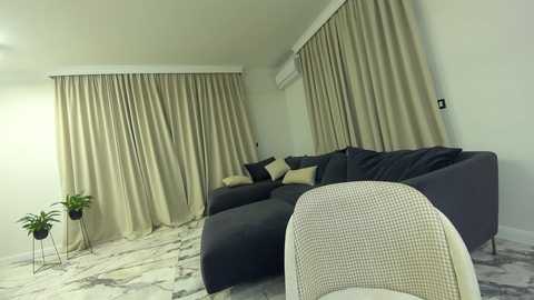 Media: Video of a modern living room with beige curtains, a gray sectional sofa, a cream chair, and a green plant on a marble-patterned floor.