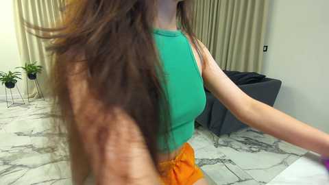 Media: Video of a woman with long brown hair in a teal sleeveless top and orange skirt, standing beside a grey chair in a room with beige curtains and marble-patterned floor.