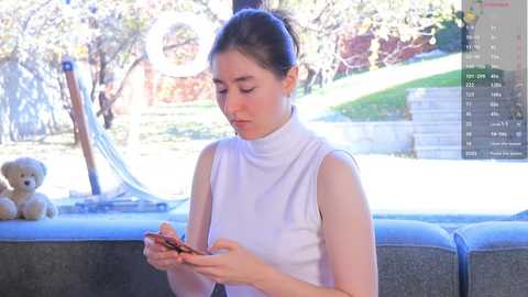 Media: Video of a young woman with fair skin, brown hair in a bun, wearing a white sleeveless turtleneck, reading a book indoors on a gray couch. Background features a window with a garden view, a teddy bear, and a digital clock display.