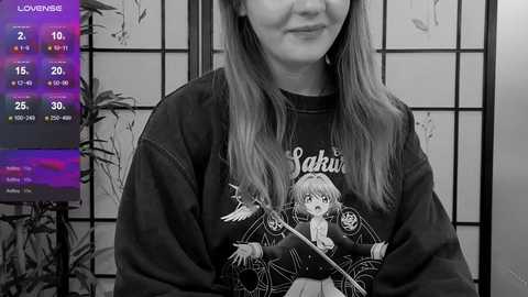 Media: Black-and-white video of a smiling woman with long hair, wearing a \"Lalisa\" graphic sweater. In the background, a shoji screen and a TV displaying a \"Love Live!\" screen.