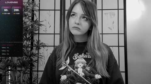 Media: Black-and-white video of a young woman with long hair, wearing a \"Sailor Moon\" T-shirt, standing in a room with a shoji screen and a potted plant, looking thoughtful.