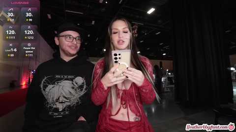 Media: Video of a man in glasses and a beanie, standing next to a woman with long blonde hair, both taking a selfie. They are in a dimly lit, industrial setting with live streaming statistics in the background.