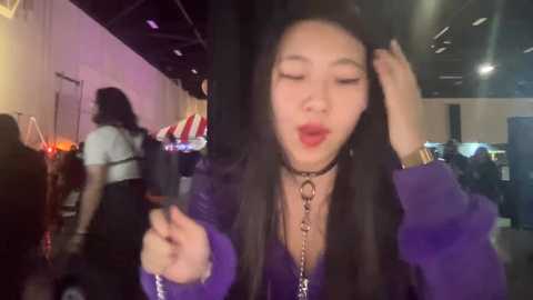 Media: Video of an East Asian woman with long black hair, wearing a purple jacket and choker, striking a playful pose indoors.