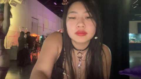 Media: Video of a young Asian woman with long black hair and red lipstick, wearing a black choker and lace top, in a dimly lit, industrial-themed venue with neon lights.