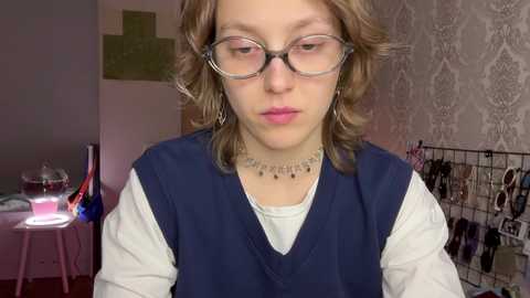 Media: Video of a young woman with shoulder-length wavy blonde hair, wearing glasses, a white t-shirt under a dark blue vest, and a necklace, standing in a room with floral wallpaper, a small table, and a wire rack.