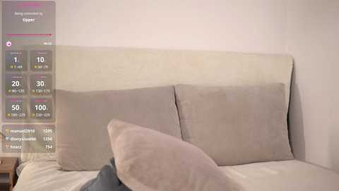 Media: Video of a minimalist bedroom with beige walls and a cream-colored upholstered headboard. The bed features two large pillows and a folded gray blanket, with a digital overlay showing weather and temperature information.