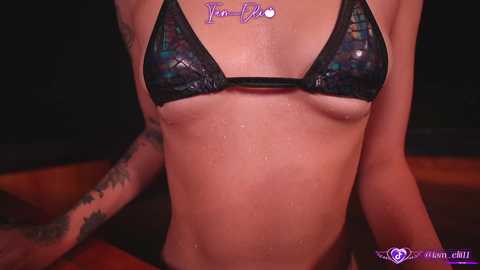 Media: Video of a slender, light-skinned woman wearing a shiny, black, sequin bikini top, revealing small breasts, and tattoos on her arms, in a dimly-lit room.