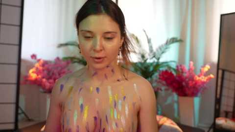 Media: Video of a fair-skinned woman with dark hair, nude, covered in colorful paint, standing in a brightly lit room with pink flowers and white curtains.