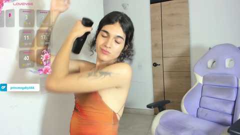 Media: Video of a young woman with curly black hair, medium skin tone, and a slim build, wearing an orange strapless dress, playing a video game on a black controller in a modern, minimalistic room with a purple gaming chair and a wooden door.