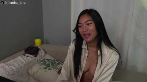 Media: Video of a smiling Asian woman with long black hair, wearing an open white shirt, sitting on a beige couch in a dimly lit living room.