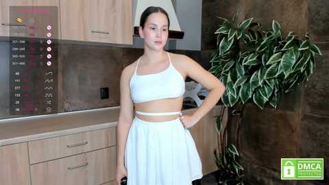 Media: Video of a young woman with fair skin and dark hair, wearing a white crop top and skirt, standing in a modern kitchen with light wooden cabinets, a stove, and a green potted plant.