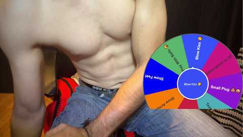 Media: Video of a shirtless, muscular, fair-skinned man sitting on a bed with blue jeans. He holds a colorful spinner wheel with game options like \"Spin to Win\" and \"Spin to Play.\" Background includes dark curtains and a red and black blanket.