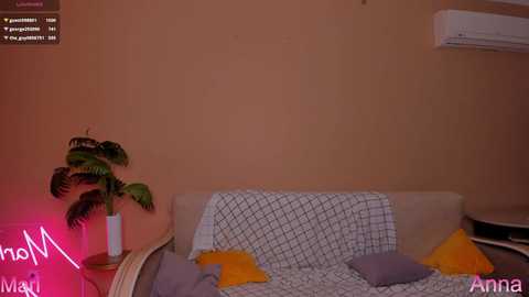 Media: Video of a cozy, minimalist bedroom with a beige wall, a bed with a white geometric-patterned blanket, two pillows in yellow and gray, a potted plant on a wooden side table, and a pink neon sign on the left.