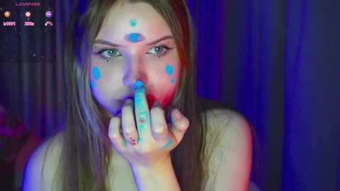 Media: A video of a young woman with long brown hair and blue face paint dots, holding her fingers to her mouth, against a dark blue background.