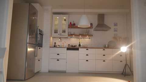 Media: Video of a modern, minimalist kitchen with white cabinets, stainless steel appliances, and a large, round pendant light; light wood flooring and a tripod floor lamp.