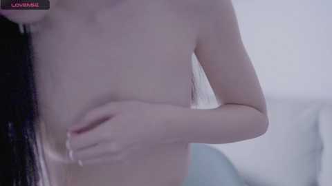 Media: Video of a light-skinned person with long black hair, topless, covering their chest with one hand, standing in a softly lit room with white walls and a bed visible in the background.