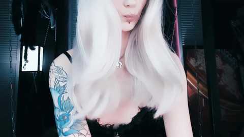 Media: Video of a pale-skinned woman with platinum blonde hair, wearing a black lace top, showcasing a blue floral tattoo sleeve and a silver necklace.