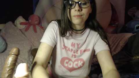 Media: A video of a young woman with long dark hair, wearing glasses and a \"MY STAPLE GUN IS A COCK\" t-shirt, holding a dildo, in a dimly-lit bedroom with plush toys.