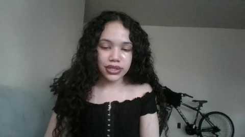 Media: Video of a young woman with curly dark hair, wearing a black off-shoulder top, standing in a simple room with a bicycle and a sofa in the background.