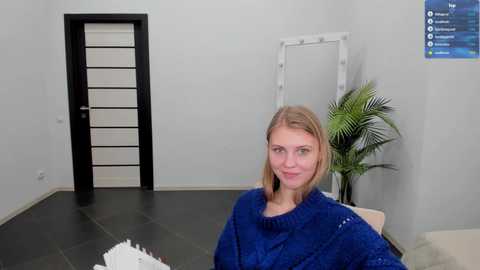 Media: A video of a blonde woman in a blue sweater sitting in a minimalist, modern room with a tall potted plant, white-framed mirror, and black door with white stripes.