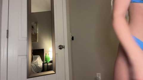 Media: Video of a bedroom with a white door and a mirror reflecting a bed, nightstand, and green lamp. A person in a blue bra stands in the foreground, partially out of focus.