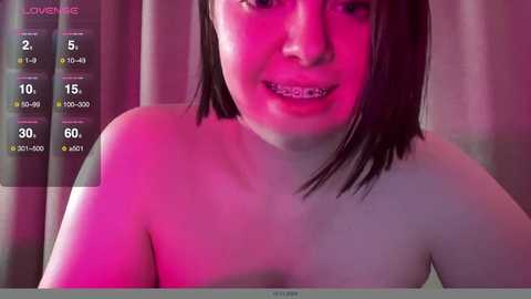 Media: A video of a topless woman with straight, shoulder-length brown hair, wearing braces, and sitting against a pink-lit background.