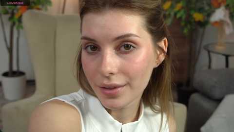 Media: Video of a young Caucasian woman with fair skin, brown hair, and a light complexion. She wears a white sleeveless top. Background features beige furniture, potted plants, and a gray couch.