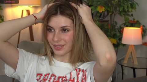 Media: Video of a young woman with fair skin and straight brown hair, wearing a white \"Raiders\" t-shirt, combing her hair in a living room with a lamp, plants, and a wooden table.